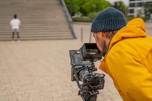 Ein Mann in gelber Jacke und blauer Mütze bedient im Freien eine professionelle Videokamera.