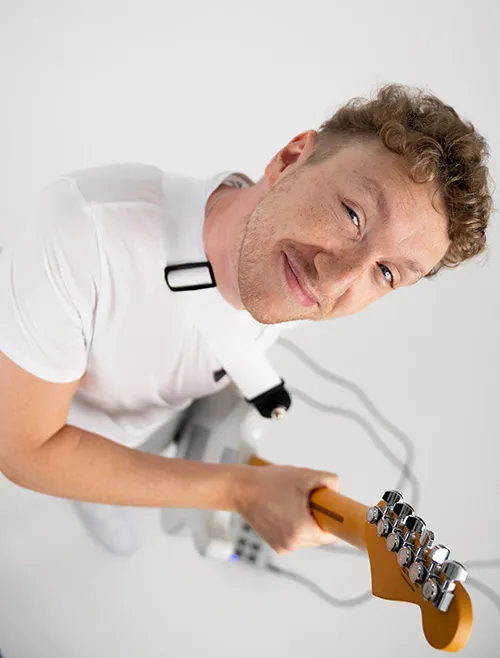 Ein lächelnder junger Mann mit lockigem Haar, der ein weißes T-Shirt trägt und eine E-Gitarre mit Gurt spielt.