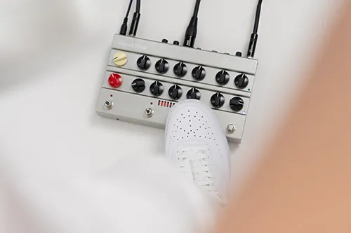 Ein Fuß in einem weißen Turnschuh drückt einen Knopf an einem grauen Gitarreneffektpedal mit mehreren schwarzen und roten Reglern und angeschlossenen Kabeln.