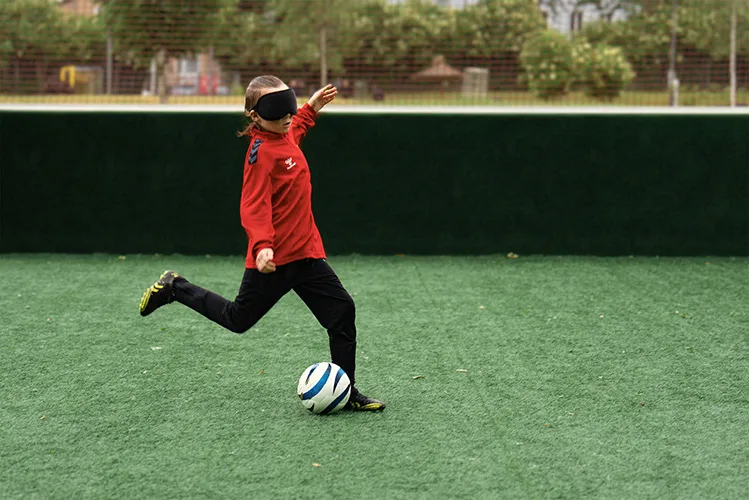 Ein Kind mit verbundenen Augen und roter Jacke kickt einen Fußball auf einem Rasenfeld.