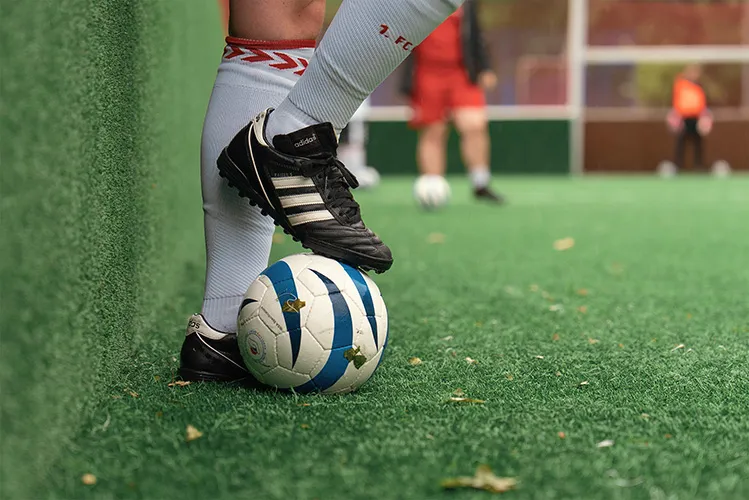 Nahaufnahme des Fußes eines Fußballspielers mit schwarzen Adidas-Schuhen, der auf einem blau-weißen Fußball auf Kunstrasen in der Nähe einer grünen Wand ruht.