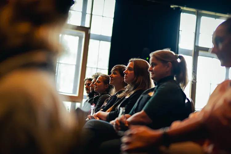 Eine Reihe von Menschen sitzt aufmerksam in einem Raum mit großen Fenstern und hört einem Redner zu.
