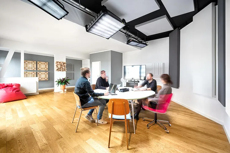 Vier Personen sitzen und arbeiten um einen weißen runden Tisch in einem geräumigen, modernen Studioraum mit Holzboden und großen Deckenleuchten.