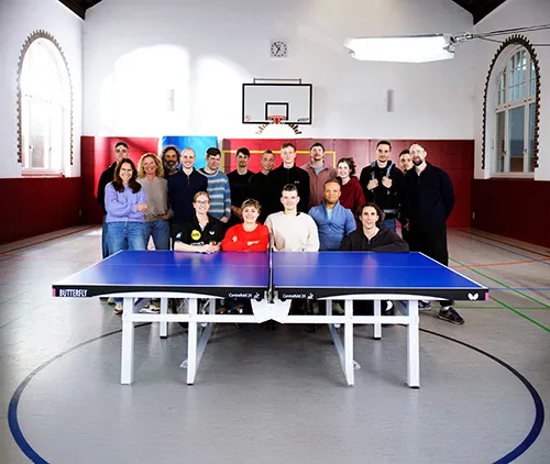 Eine Gruppe von 19 Personen posiert hinter einer blauen Tischtennisplatte in einer Sporthalle mit Basketballkorb.