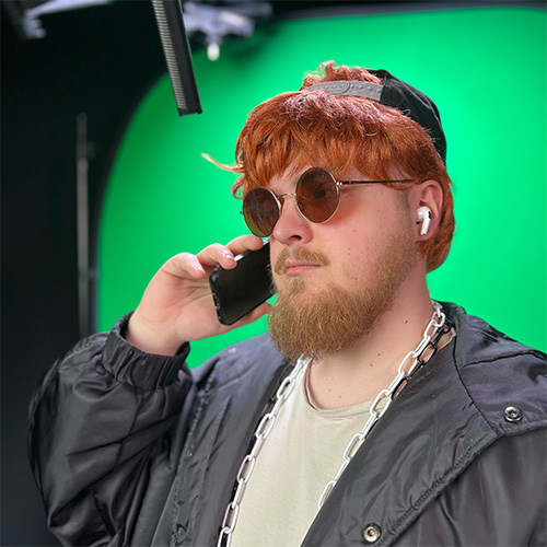 Junger Mann mit rotem Haar, Sonnenbrille und Kapuzenjacke telefoniert mit Smartphone vor grünem Hintergrund.