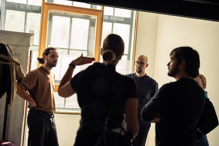 Fünf Personen im Gespräch in einem Raum mit großen Fenstern, eine Frau im Vordergrund gestikuliert.