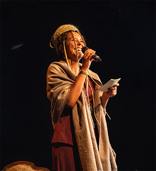 Frau mit geflochtenen Haaren spricht mit Mikrofon und Zettel vor schwarzem Hintergrund.