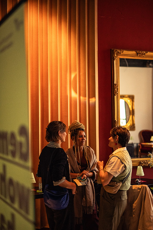 Drei Personen unterhalten sich vor einem goldgerahmten Spiegel in einem warm beleuchteten Raum mit roten Wänden und gelben Vorhängen.