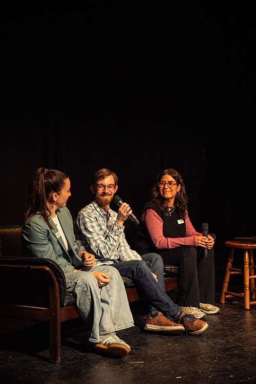 Drei Personen sitzen auf einem Sofa auf einer Bühne und sprechen in Mikrofone, umgeben von dunklem Hintergrund.