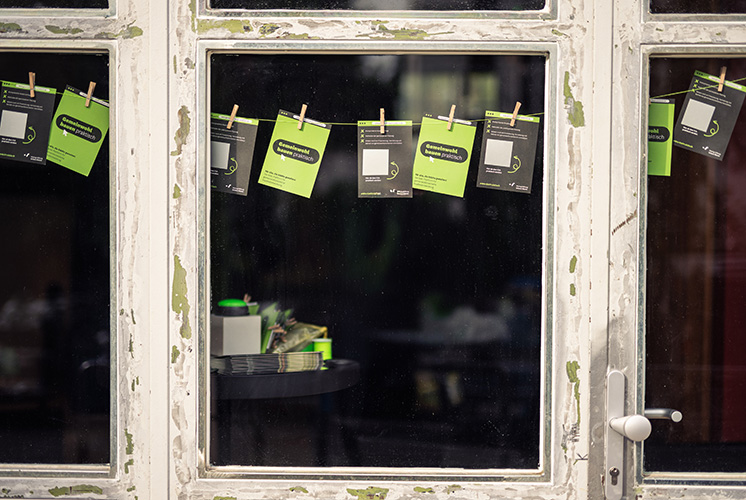 Verwittertes weißes Fenster mit Klammern, die grüne und schwarze Informationskarten halten, vor dunklem Hintergrund und einem Tisch mit gestapelten Karten.