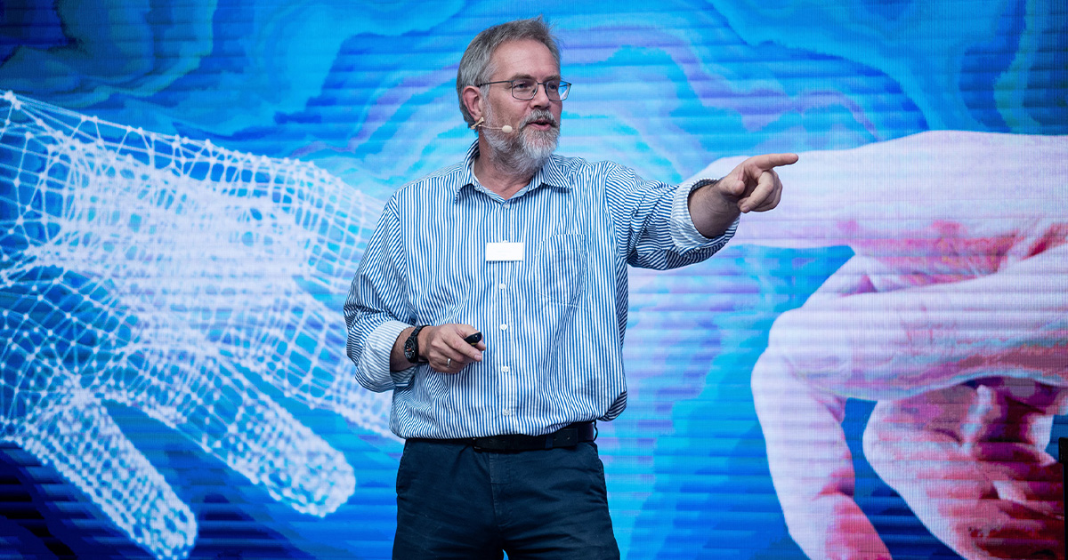 Ein älterer Mann mit Brille und Mikrofon-Headset spricht und zeigt nach rechts vor einem Bildschirm mit zwei Händen, eine digital und eine real dargestellt.