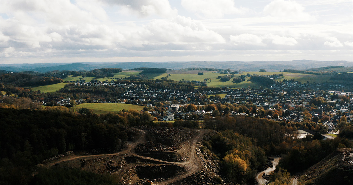Landschaft mit Wald, einem Baugebiet im Vordergrund, Feldern und einem weitläufigen Dorf unter bewölktem Himmel.