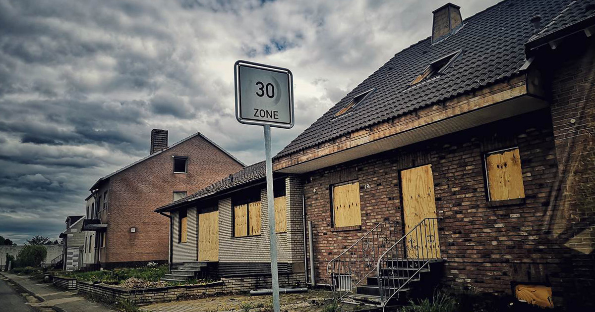 Verlassene Häuser mit vernagelten Fenstern und Türen an einer Straße mit einem 30-Zone-Verkehrsschild unter bewölktem Himmel.
