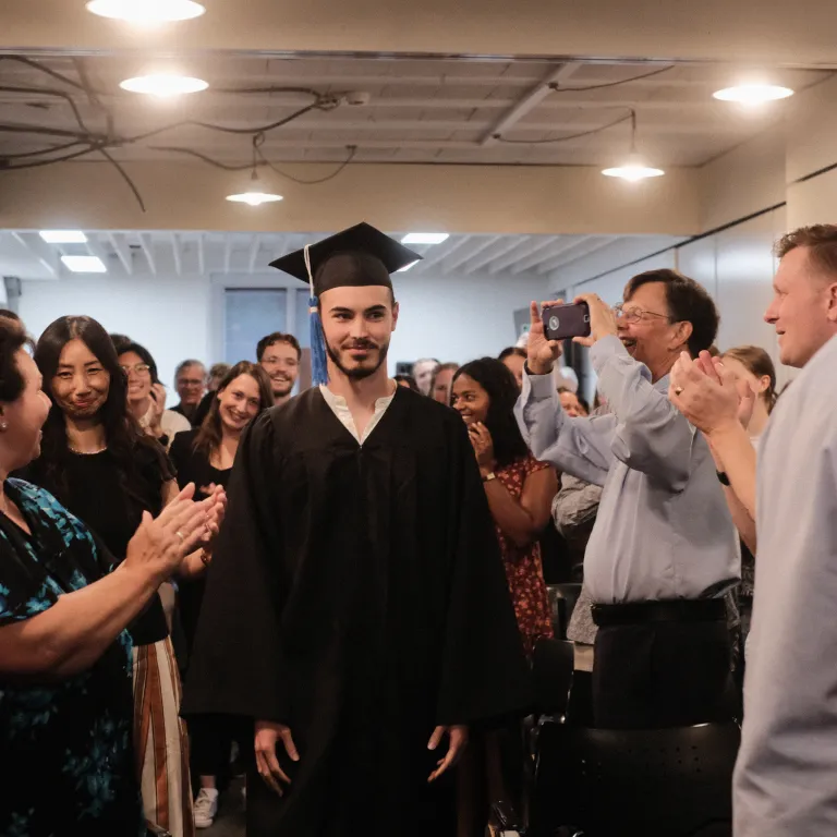 A man clothed in a graduation gown and cap walking through a crowd of clapping people.
