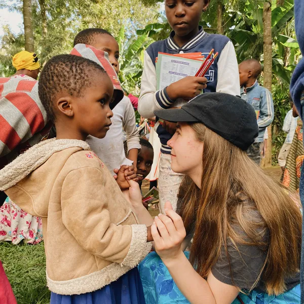 A female student kneeling down, smiling, and holding the hands of a young child.