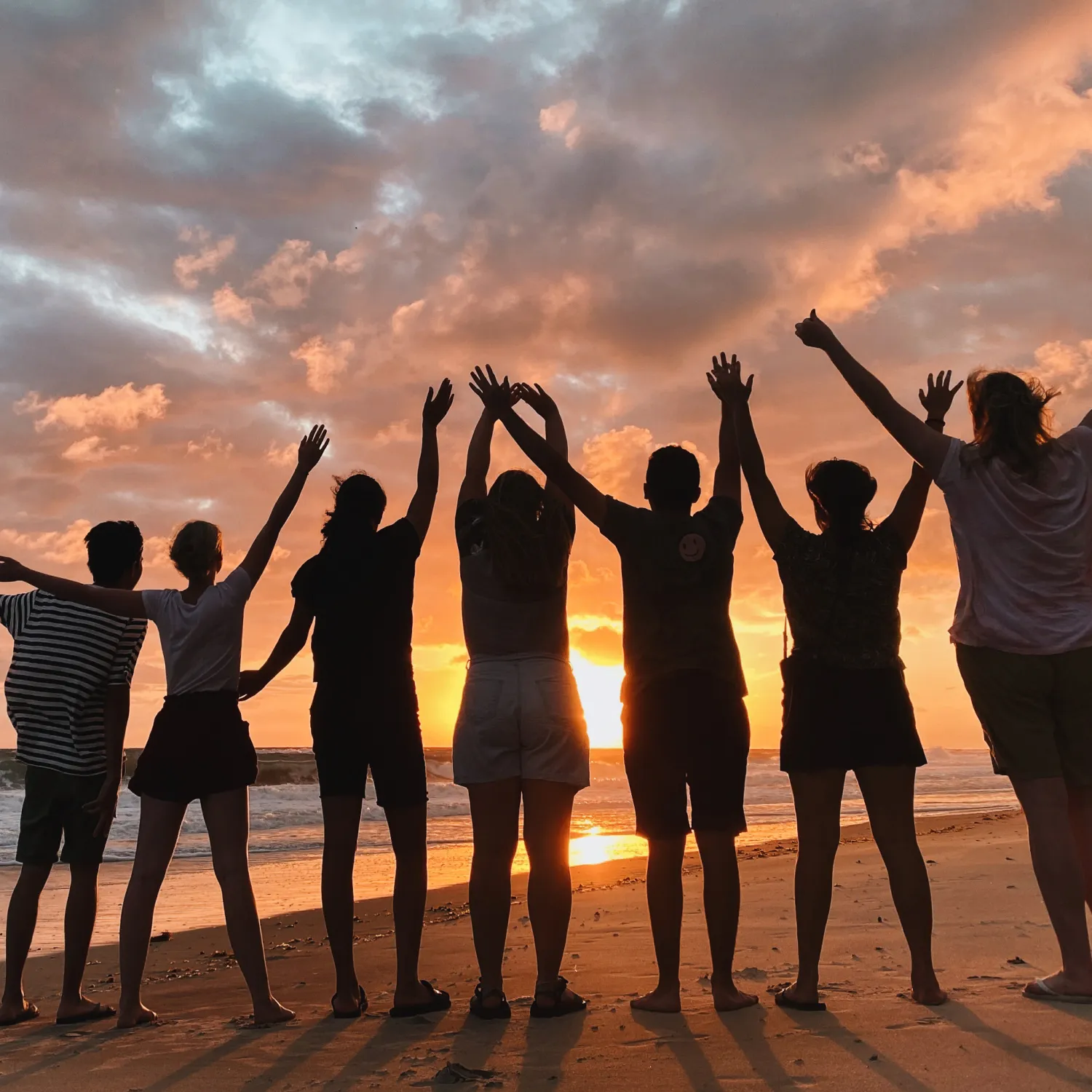A group of students with linked arms up in the air standing on the beach in front of the ocean with the sun setting.