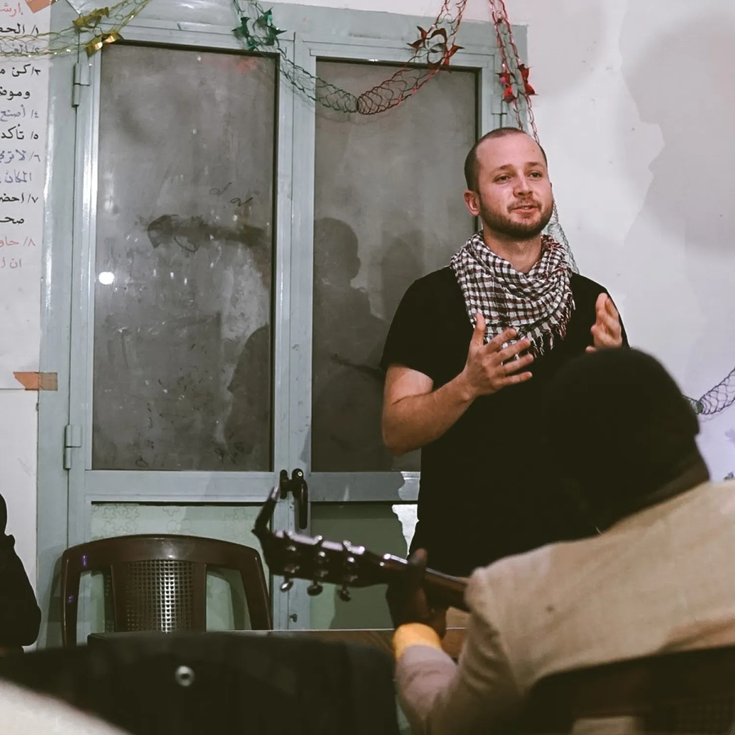 A man standing in front of a door, wearing a scarf, and speaking with his hands lifted in front of him.