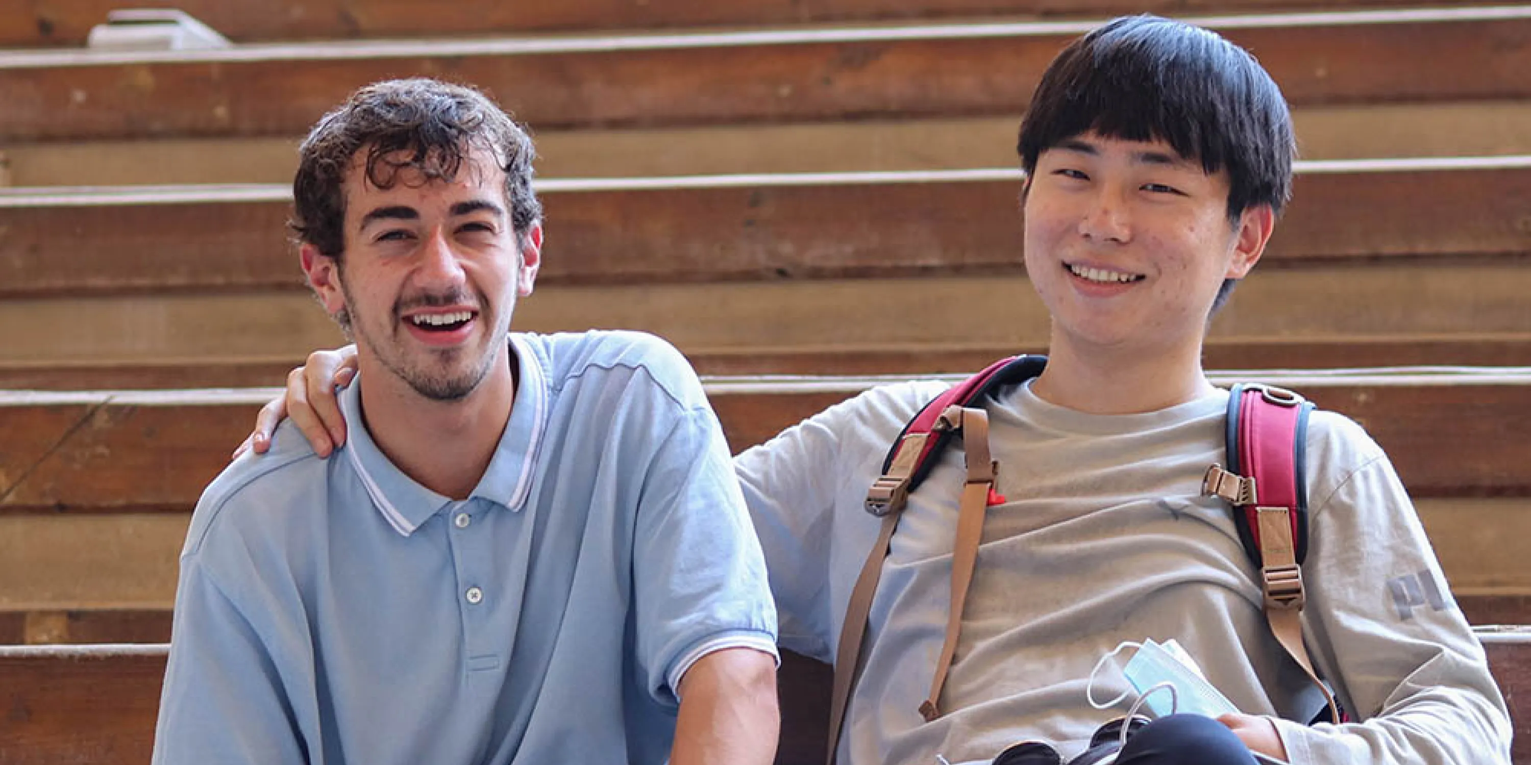 Two male students seated on stairs and smiling with one placing his arm over the shoulder of the other.