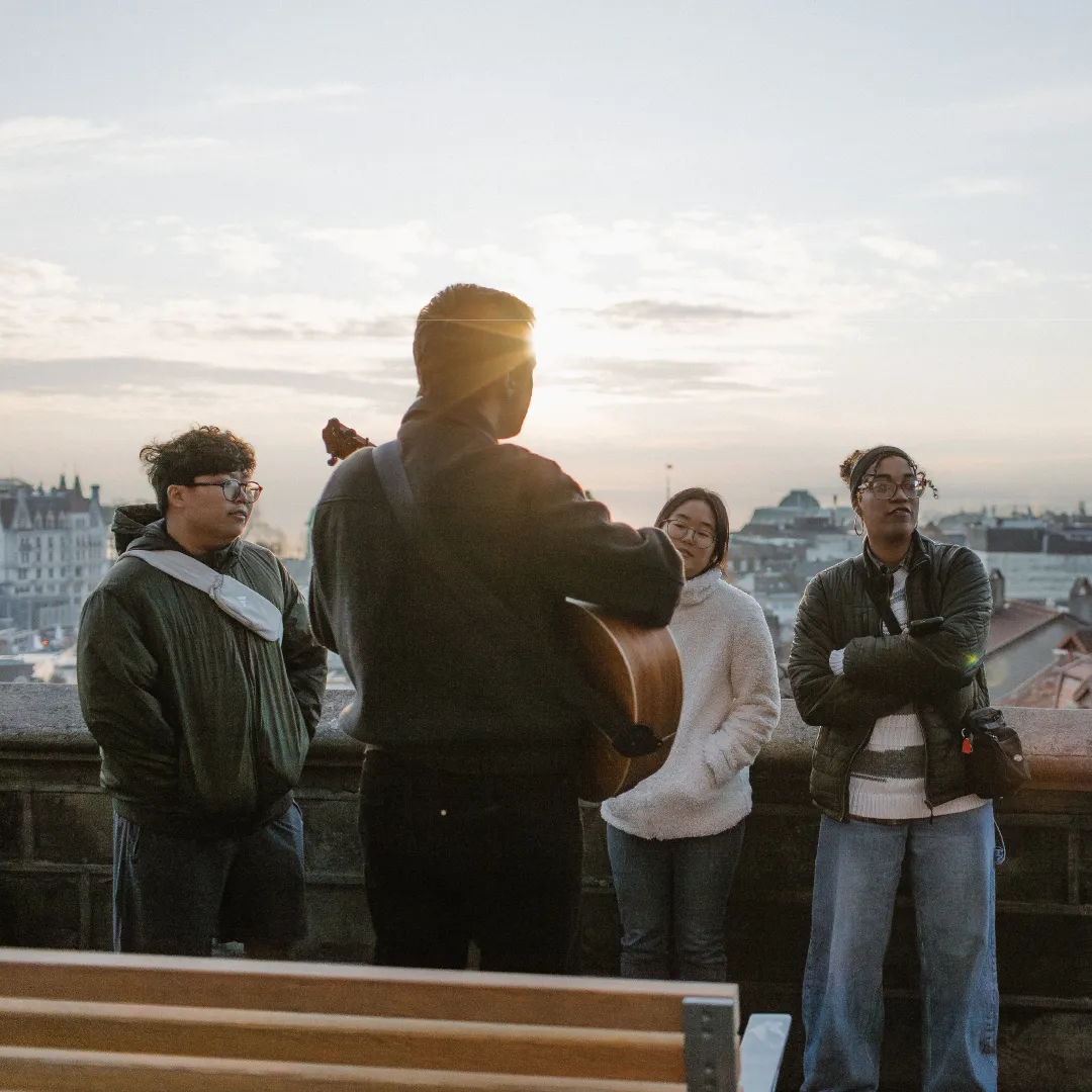 Person playing guitar to a group of three people standing by a stone railing at sunset with cityscape in the background.