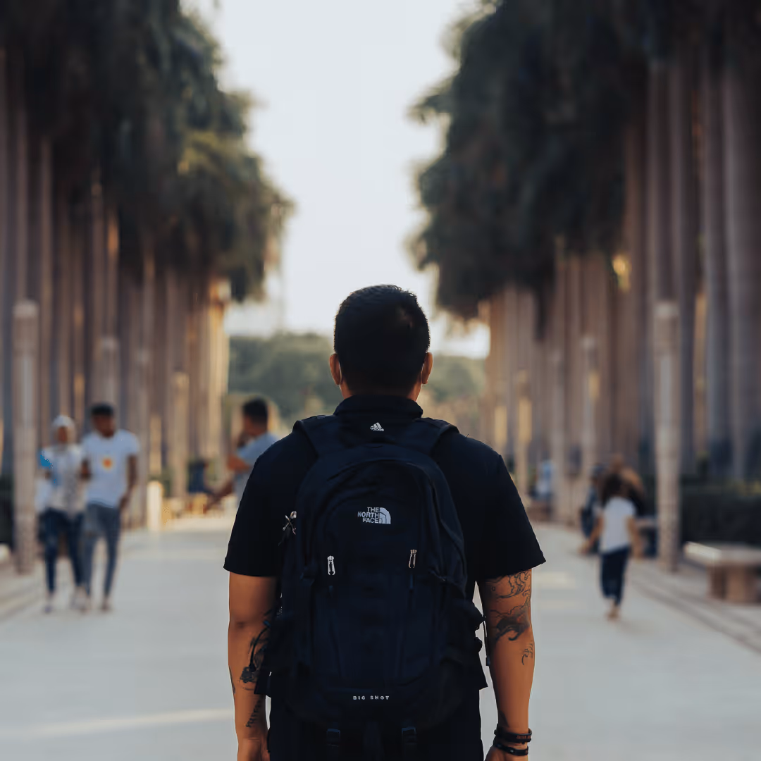 Man with tattoos wearing a black backpack walking down a palm-lined urban walkway with blurred people in the background.