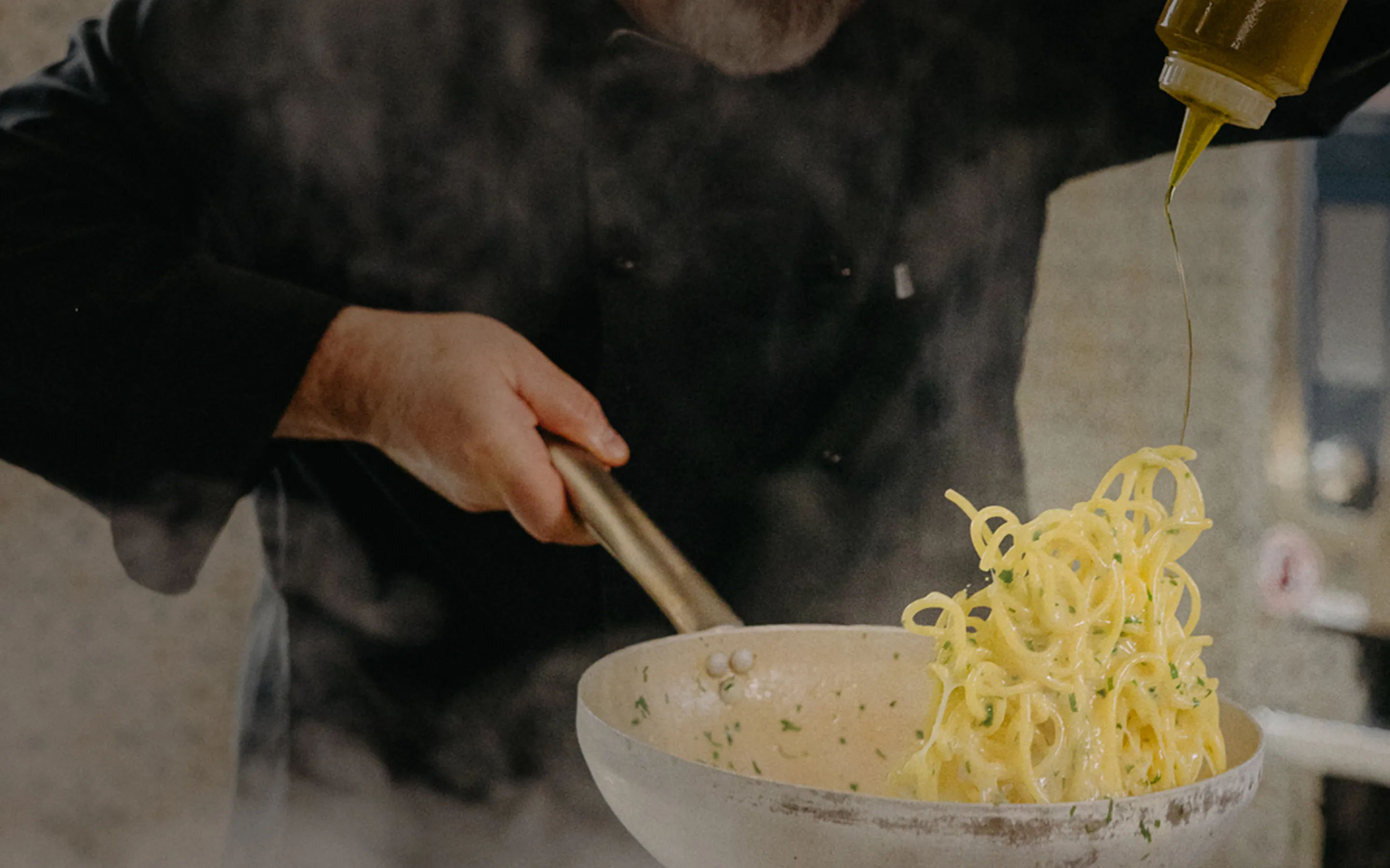 Chef tossing spaghetti in a pan while drizzling olive oil.
