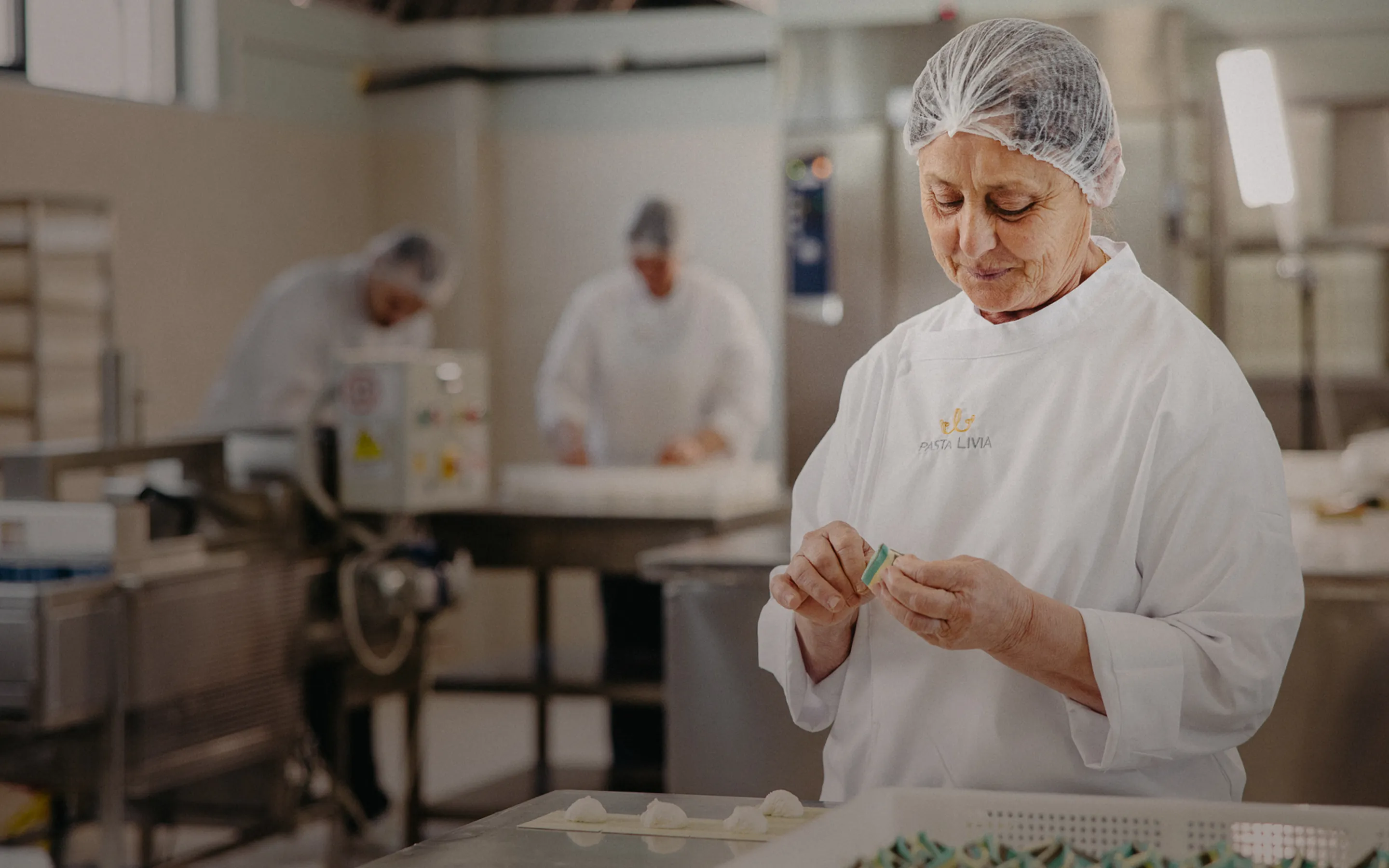 Woman in white Pasta Livia uniform and hairnet handcrafting pasta in a clean kitchen environment.