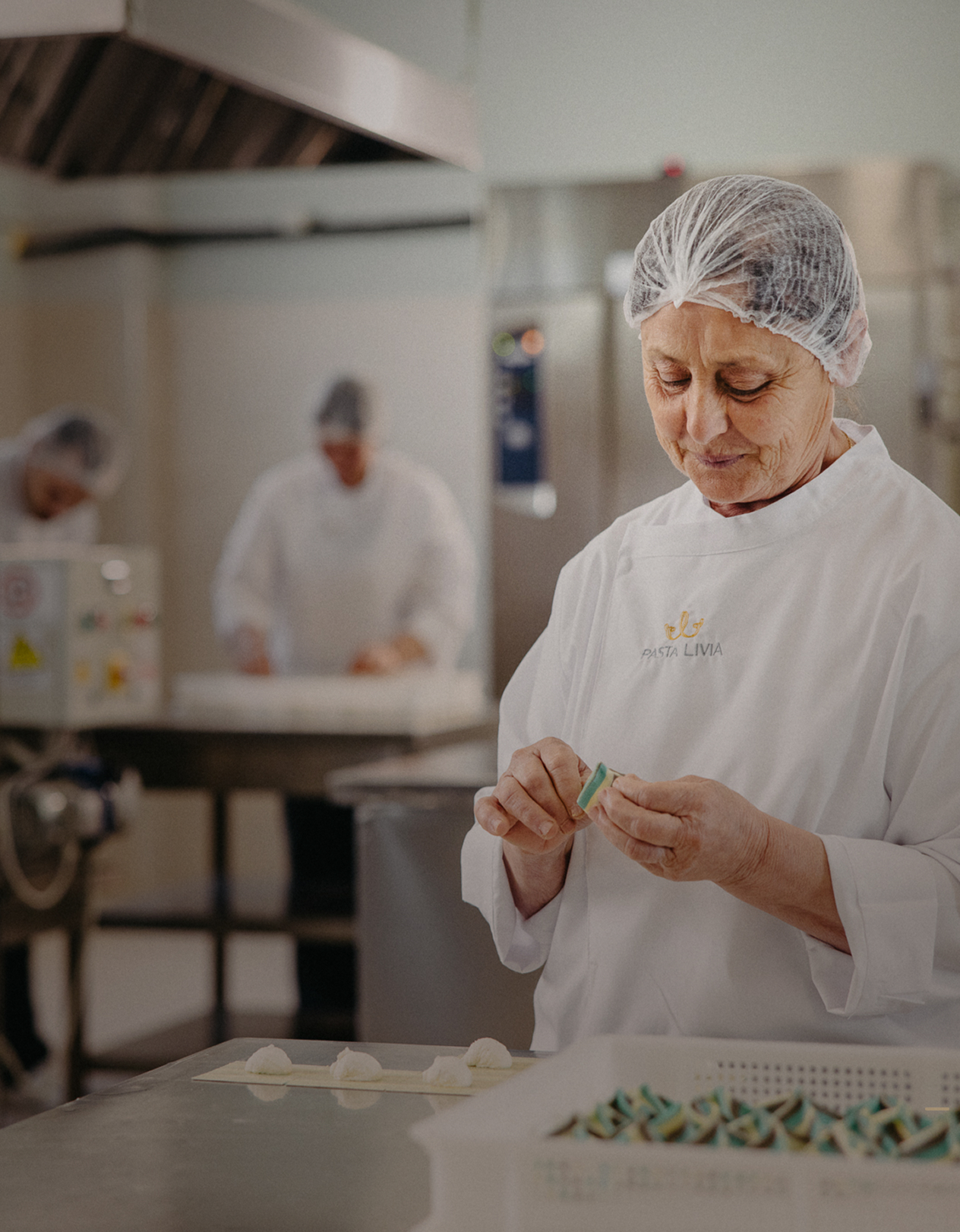 Woman in white Pasta Livia uniform and hairnet handcrafting pasta in a clean kitchen environment.
