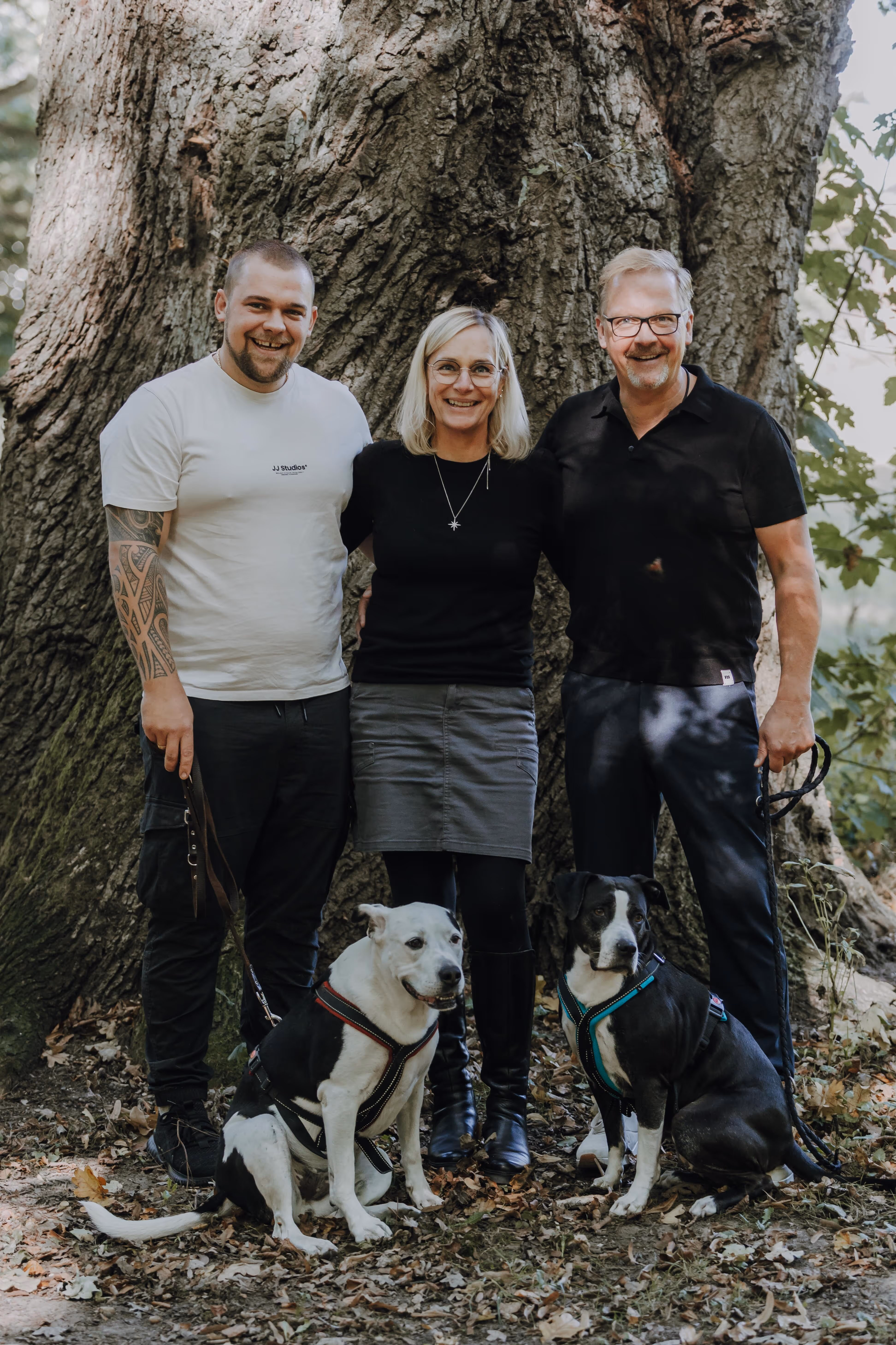 Familie Faasch, zwei Männer und eine Frau, stehen vor einem großen Baum mit zwei angeleinten Hunden vor ihnen.