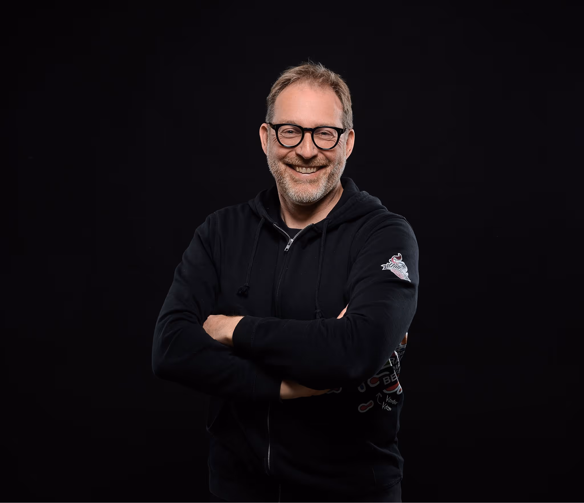 Mann mit Brille und schwarzem Kapuzenpulli lächelt vor schwarzem Hintergrund mit verschränkten Armen.