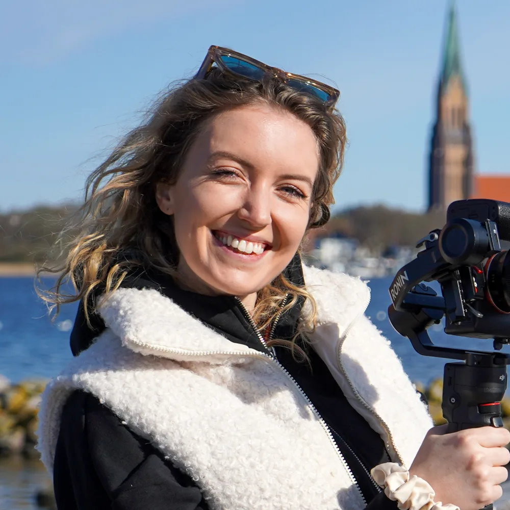 Lächelnde Frau mit lockigem Haar hält eine Kamera mit Gimbal vor einem Gewässer.