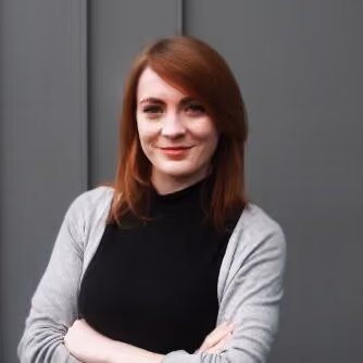 Smiling woman with red hair wearing a black top and gray cardigan against a gray background.
