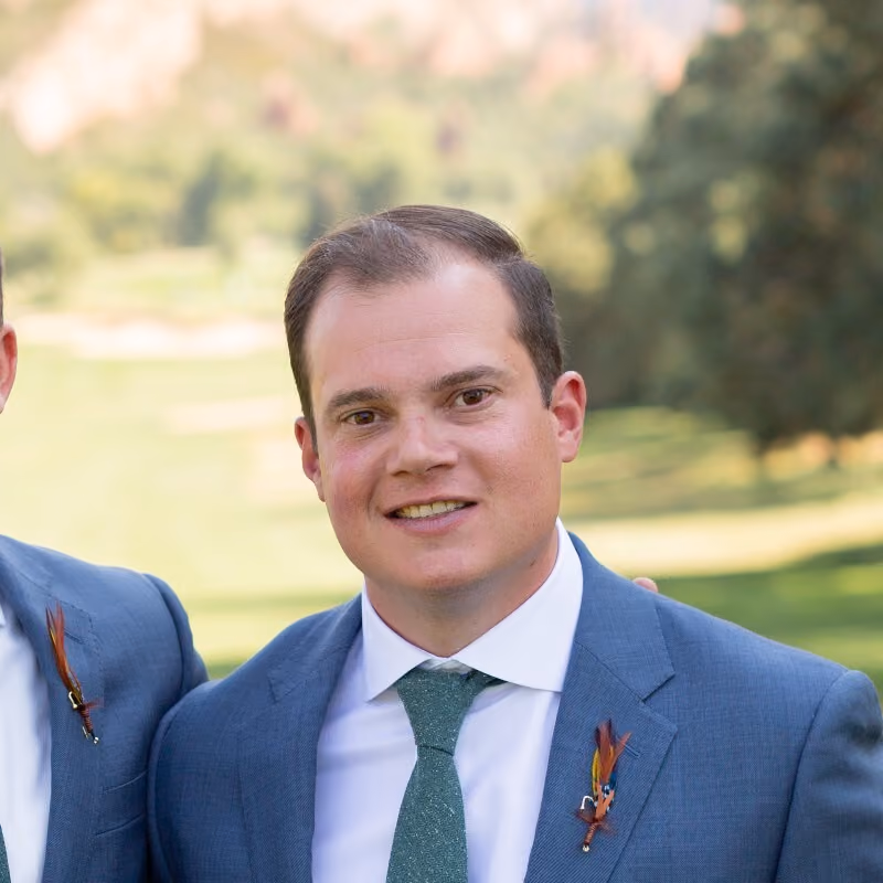 Smiling man in a blue suit and green tie with a feather boutonniere outdoors in a blurred green background.