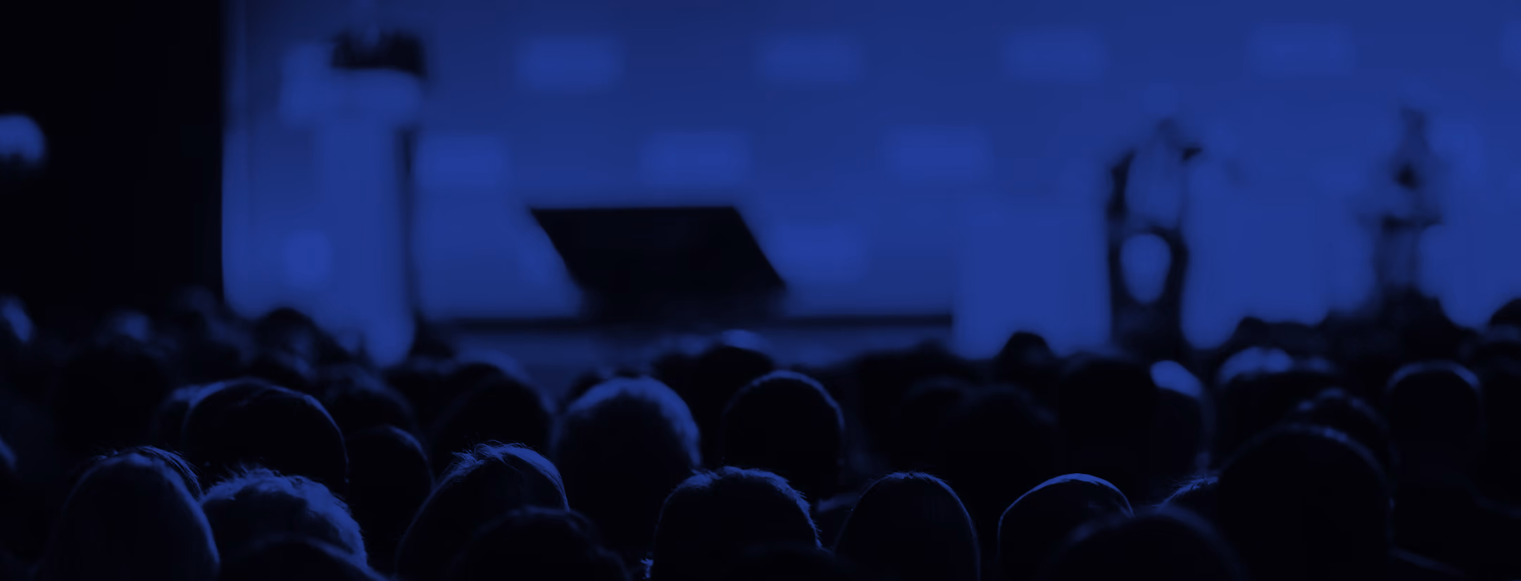 Audience watching a blurred stage with two speakers during a dark blue-lit event.