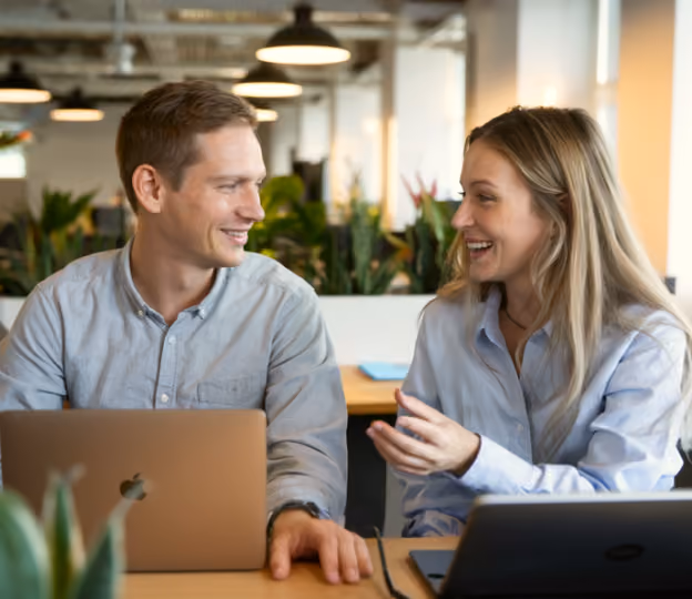 Smiling man and woman sitting at a desk with laptops, engaged in a friendly conversation.