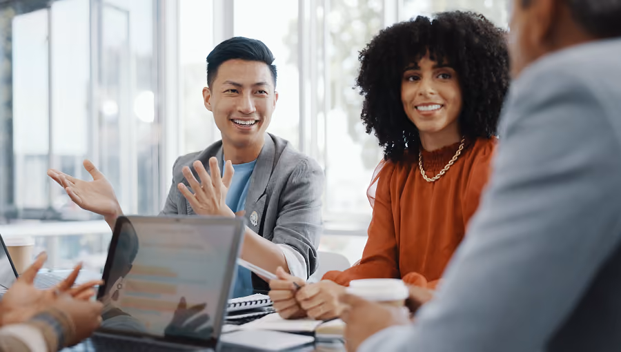 Two colleagues smiling and talking during a meeting in a bright office with laptops and coffee cups.