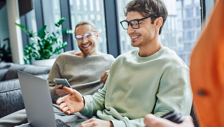Two men sitting on a couch laughing while using a laptop and smartphone in a bright modern office.