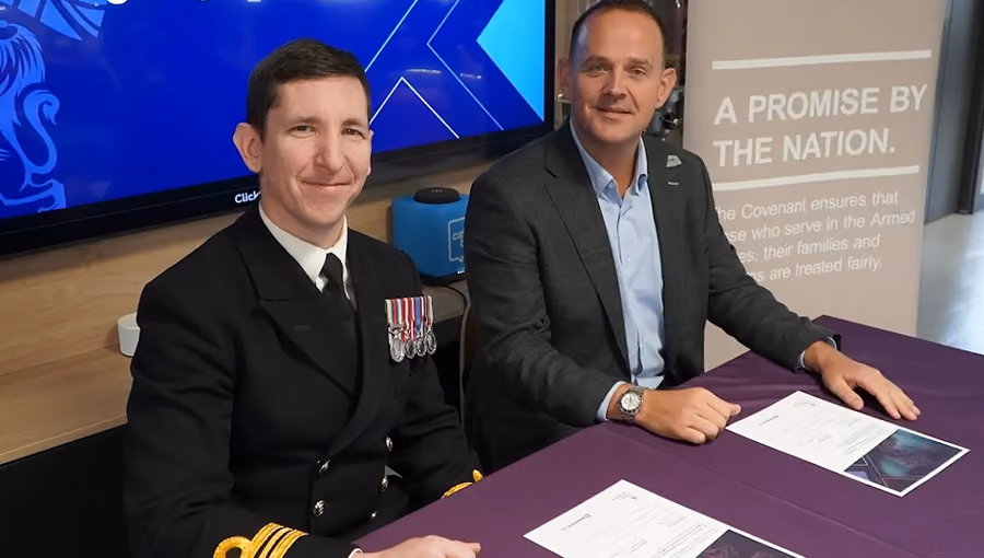 Two men seated at a table signing documents; one wears a military uniform with medals, the other a suit.