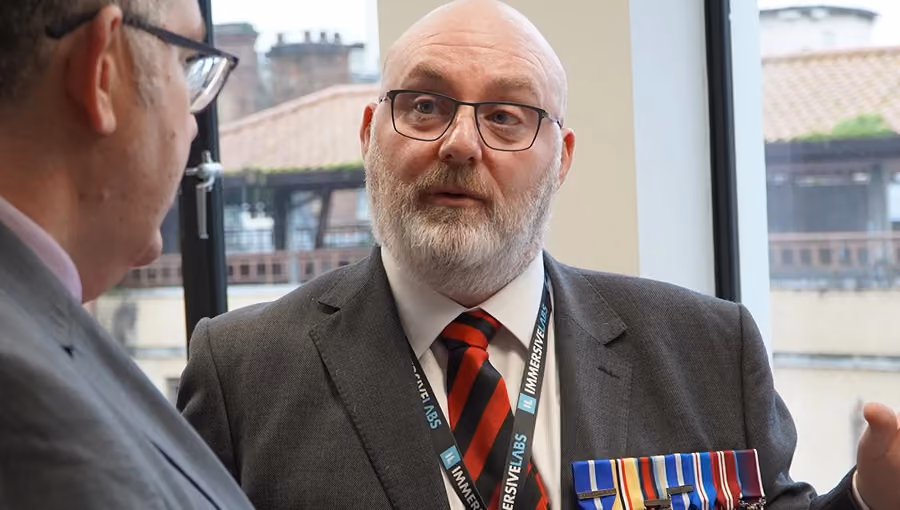 Bearded man in glasses and suit with military medals talking to another man indoors near a window.