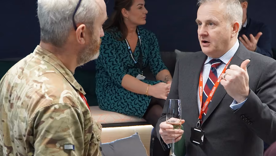 Man in camouflage military uniform talks with man in suit holding a glass and gesturing thumbs up.