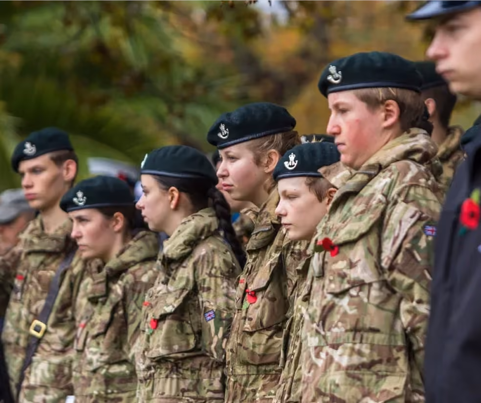 Line of young soldiers in camouflage uniforms and berets with red poppies standing outdoors.