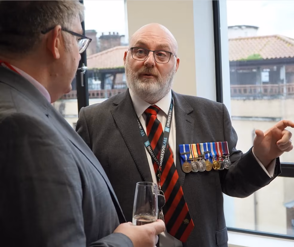Two men in suits having a conversation, one wearing several medals and holding a glass of drink.