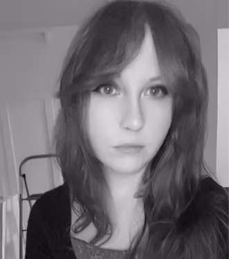 Black and white portrait of a woman with long hair and bangs, looking directly at the camera with a neutral expression, indoors with a partially visible door and furniture in the background.