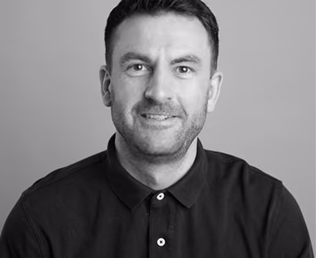 Black and white portrait of a man with short dark hair and light facial hair, wearing a black collared shirt, smiling gently at the camera with a plain background.