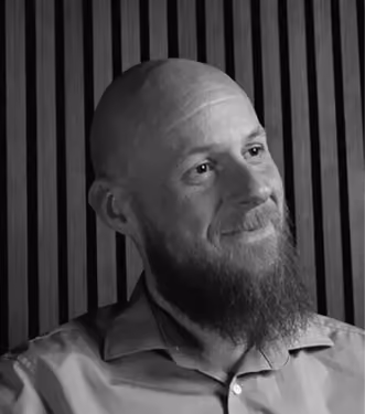 Black and white portrait of a bald man with a beard, wearing a collared shirt, sitting in front of a vertical striped wooden wall, looking slightly to the right with a gentle smile.