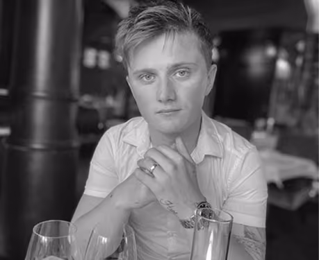 Black and white photo of a young person with short hair wearing a short-sleeve shirt, sitting at a table with hands clasped and looking directly at the camera, with glasses and a watch visible on the table.