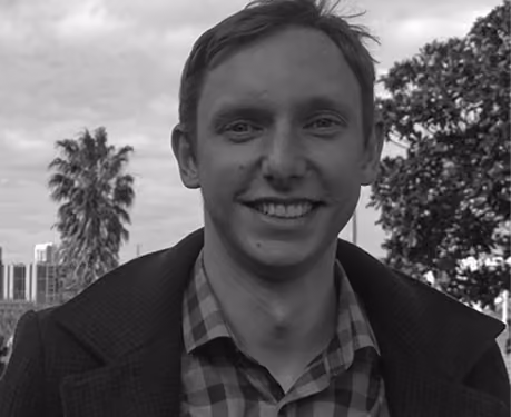 Black and white photo of a smiling young man wearing a checkered shirt and dark jacket, outdoors with trees and buildings in the background.