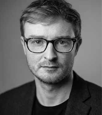 Black and white portrait of a man with short hair and glasses wearing a dark jacket over a dark shirt, looking directly at the camera against a plain background.