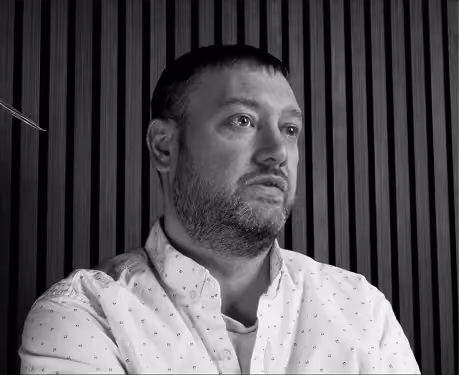 Black-and-white photo of a man with short hair and a beard wearing a light-colored shirt with small patterns, looking to the side against a vertical striped wall background.