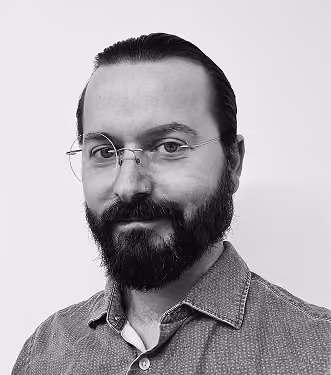 Black and white portrait of a man with a full beard, mustache, and glasses, wearing a buttoned-up patterned shirt, looking at the camera with a slight smile.