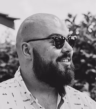 Bearded man with a bald head wearing dark sunglasses and a light-colored shirt with a small pattern, smiling and looking to the right with blurred trees in the background.
